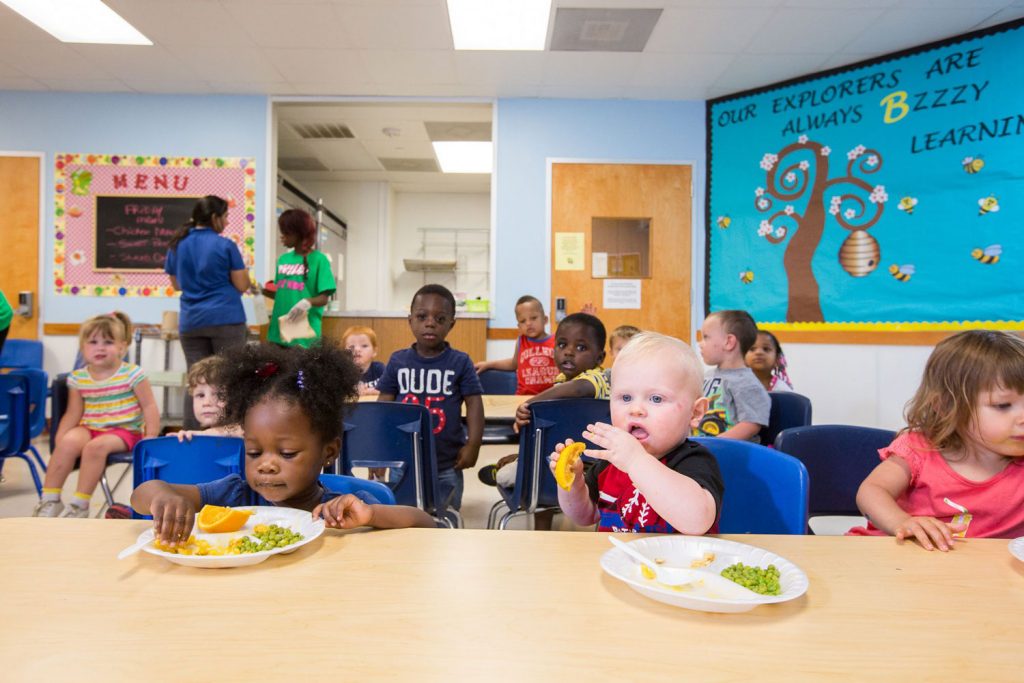 Children sit at a table eating snacks while others sit on the floor with caregivers in a brightly decorated classroom, exemplifying the nurturing environment of this early education franchise.