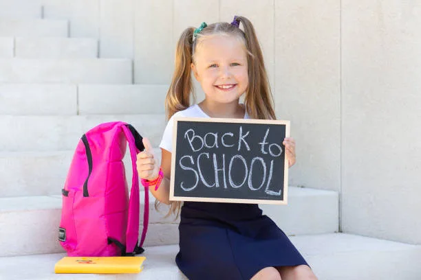 A young girl with pigtails sits on outdoor steps holding a "Back to School" sign. She is smiling, with a pink backpack and yellow book beside her, ready for the exciting journey ahead at her early childhood education franchise.