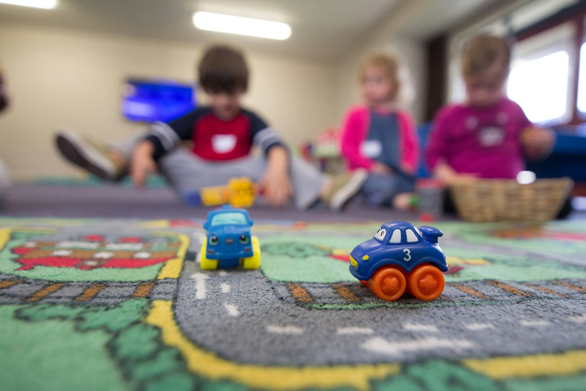 Toy cars on a road-patterned classroom carpet with children playing in the background.